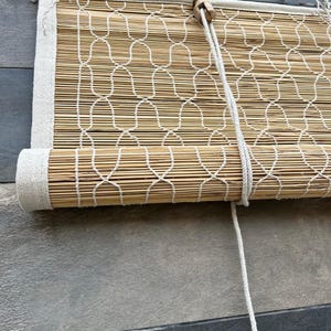 May include: A roll-up window shade constructed from thin, light brown bamboo slats. The shade features a white woven cord pattern overlaid on the bamboo and is finished with a thick white fabric border along the edges. A white pull cord is attached.