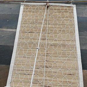 May include: A rectangular bamboo window shade with a white patterned design. The shade is framed with a white fabric border and has a rolled bottom. A white cord hangs from the top, connected to a wooden toggle.