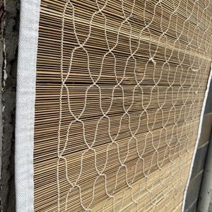 May include: A rectangular bamboo blind featuring thin, horizontal light brown wooden slats held together by a white woven cord net overlay. The entire structure is framed by a thick, white fabric border along the vertical and horizontal edges.