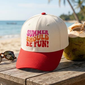 May include: A baseball cap with a cream-coloured crown and a red peak. The cap has the embroidered text "SUMMER SHOULD BE FUN!" in pink and orange. A red button is on top. The background shows a beach scene and a coconut.