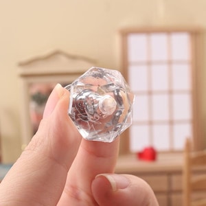 May include: Clear, faceted crystal cabinet knob with a silver-toned base. The knob has an octagonal shape and is held in a hand. The background is blurred, suggesting a room setting.
