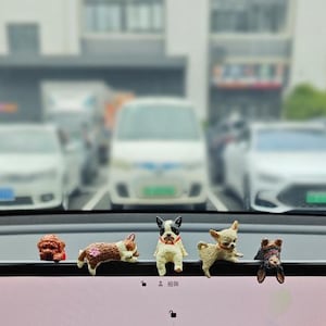 May include: A collection of small dog figurines in various breeds and poses, arranged on a car dashboard. The dogs are painted in realistic colors and details. The background shows a blurred street scene with parked cars.
