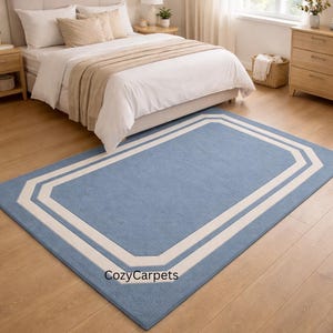 May include: A rectangular blue rug with a white border in a bedroom. The rug features a light blue centre and a white geometric border. It's placed on a wooden floor in front of a bed.