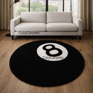 May include: A circular black rug featuring a white number eight design, reminiscent of a snooker ball. The rug is positioned on a wooden floor, in front of a light-coloured sofa. The text "CozyCarpets" is printed on the rug.