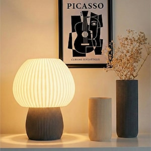 May include: A modern table lamp with a ribbed, mushroom-shaped shade and a gray base. Two ribbed vases, one beige and one dark gray, stand beside the lamp. A framed black and white Picasso print hangs in the background.