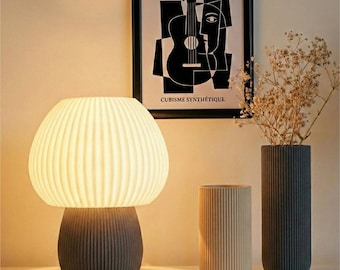 May include: A modern table lamp with a ribbed, mushroom-shaped shade and a gray base. Two ribbed vases, one beige and one dark gray, stand beside the lamp. A framed black and white Picasso print hangs in the background.
