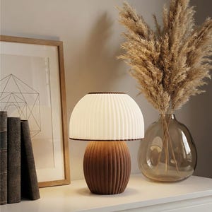 May include: A brown and cream-colored table lamp with a ribbed base and a pleated lampshade. The lamp is on a white surface, next to a framed geometric print and a vase with dried plants. The lamp is a decorative home accent.
