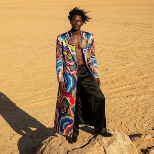 May include: A long, colourful coat with a swirling pattern of blues, greens, pinks, and yellows. The coat is worn with black trousers and shoes. The background is a desert landscape with a large rock.