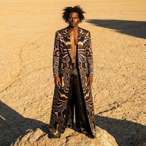 May include: A person stands on a rock in a desert landscape, wearing a long coat and black trousers. The coat features a black, brown, and rainbow-coloured swirling pattern. The coat is open, revealing the person's bare chest.