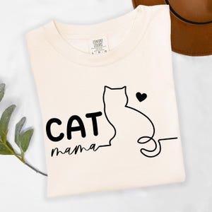 May include: Cream-colored t-shirt with the words "CAT mama" in black, along with a black cat outline and heart. The shirt is folded and has a Comfort Colors tag.