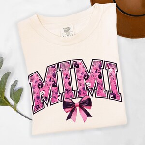 May include: Cream-colored t-shirt with the word "MIMI" in a pink and purple Halloween-themed design. The letters are filled with spiderwebs, skulls, and pumpkins. A black and pink bow is centered below the word. The shirt is folded.