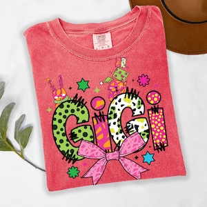 May include: Red Comfort Colors t-shirt with the name "GIGI" in a playful, colorful design. The letters are decorated with polka dots, stripes, and a pink bow. The shirt features cartoon graphics of a bunny and a dog.