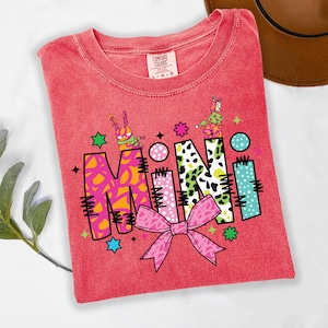 May include: A red t-shirt with the word "MINI" in a colorful, playful design. The "M" is pink and orange, the "I" is green and white, the "N" is leopard print, and the second "I" is blue. A pink bow is at the bottom.