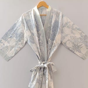 May include: A white and blue patterned dressing gown with a tie waist. The gown features a botanical design with palm trees and other foliage. The sleeves and lapels have a striped border. It hangs on a wooden hanger.