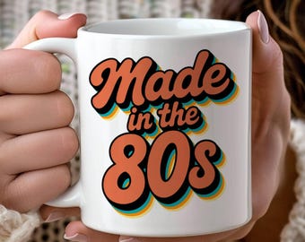 May include: White ceramic mug with the text "Made in the 80s" in orange, black, and yellow lettering. The mug has a white handle and is held by a person wearing a white sweater.