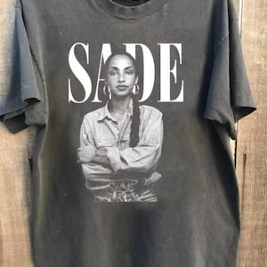 Puede incluir: Camiseta gris oscuro con una imagen en blanco y negro de Sade Adu. El nombre "SADE" está impreso en blanco sobre la imagen. Sade lleva una camisa vaquera y pendientes de aro.