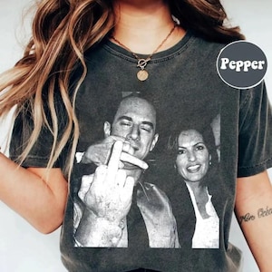 May include: Dark gray t-shirt featuring a black and white image of two people. One person is giving the middle finger. The word "Pepper" is printed on the upper right side of the shirt.