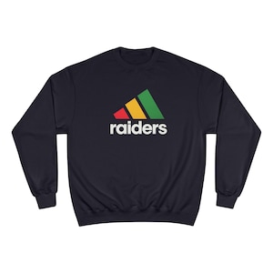 Könnte beinhalten: Marineblaues Sweatshirt mit einem farbenfrohen Logo mit roten, gelben und grünen Streifen und dem Wort "raiders" in Weiß. Das Sweatshirt hat einen Rundhalsausschnitt und lange Ärmel. Ein lässiges, bequemes Kleidungsstück.