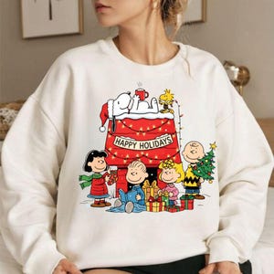 May include: A cream-coloured sweatshirt with a festive Peanuts design. Snoopy is on his doghouse, adorned with lights and the words "Happy Holidays". Other characters are gathered around gifts. A Christmas tree is held by one of the characters.