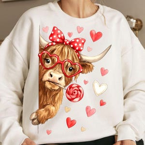 Può includere: Una felpa bianca con un'illustrazione di una mucca delle Highlands. La mucca indossa occhiali a forma di cuore, un fiocco a pois e un lecca-lecca. Cuori rossi e rosa circondano la mucca, creando un tema di San Valentino.