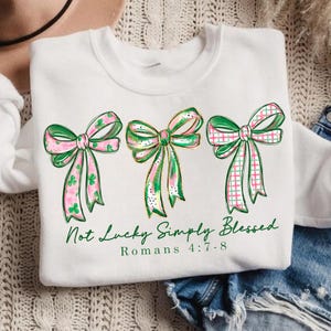May include: White sweatshirt featuring three decorative bows in green and pink with gold accents. The bows have different patterns, including shamrocks and plaid. The text "Not Lucky Simply Blessed" and "Romans 4:7-8" are printed in green.