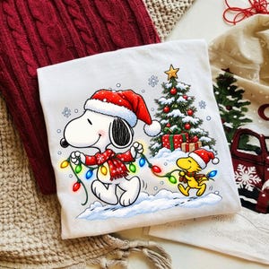 Puede incluir: Una camiseta blanca con un gráfico de Snoopy y Woodstock con gorros de Papá Noel. Snoopy sostiene una cadena de luces navideñas de colores, mientras Woodstock está cerca. Un árbol de Navidad decorado con regalos aparece de fondo.