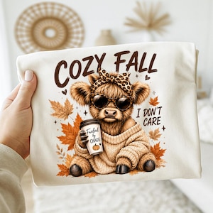 Può includere: Una t-shirt color crema con la grafica di una soffice mucca highland che indossa una fascia leopardata e occhiali da sole. La mucca indossa un maglione beige, tiene in mano un bicchiere di caffè con la scritta "Fueled by CHAOS" ed è circondata da foglie autunnali con il testo "COZY FALL I DON'T CARE."