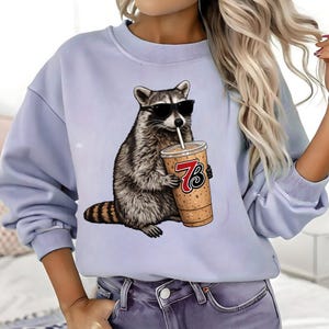 Puede incluir: Sudadera azul claro con un gráfico de un mapache con gafas de sol y sosteniendo una bebida de café con el número 73. El mapache es en tonos grises y marrones. La taza de café es de color beige con un logotipo rojo y negro.