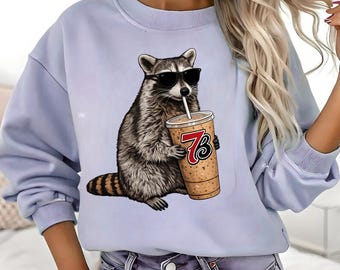 May include: A light blue sweatshirt featuring a graphic of a raccoon wearing sunglasses and holding a coffee drink with the number 73. The raccoon is in shades of gray and brown. The coffee cup is tan with a red and black logo.