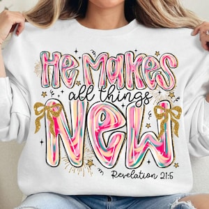 Può includere: Felpa bianca con la scritta "He Makes all things NEW" in un font colorato e a bolle. Il design include dettagli glitterati dorati, stelle e fiocchi. Il riferimento biblico "Revelation 21:5" è in basso.