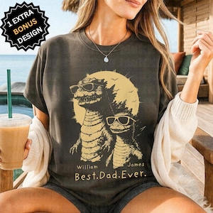 Op de afbeelding: Donkergrijs T-shirt met een afbeelding van twee dinosaurussen met zonnebrillen voor een gele zon. De tekst luidt "William James Best.Dad.Ever." Een bonusontwerp is ook inbegrepen.