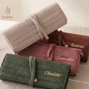 Puede incluir: Una colección de estuches rectangulares personalizados para brochas de maquillaje en varios colores: beige, burdeos, rosa y verde. Cada estuche está hecho de un material suave y texturizado y presenta una inscripción dorada con un nombre. Los estuches están atados con una correa a juego.