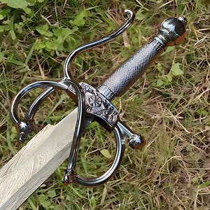 May include: A decorative sword with a silver-colored handle and guard. The blade has a unique pattern. The sword is lying on a bed of green grass.