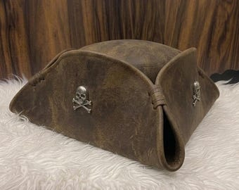 May include: A brown tricorn hat featuring a distressed leather-like texture and a silver-toned metal skull and crossbones emblem attached to the side. The hat has a wide, folded brim with stitched detailing and a rounded crown structure.