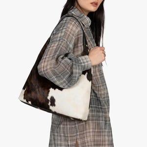 May include: A shoulder bag featuring a cowhide pattern with patches of dark brown and white fur. The bag has a structured silhouette with black leather straps and is worn over the shoulder, showcasing a natural animal print texture throughout.