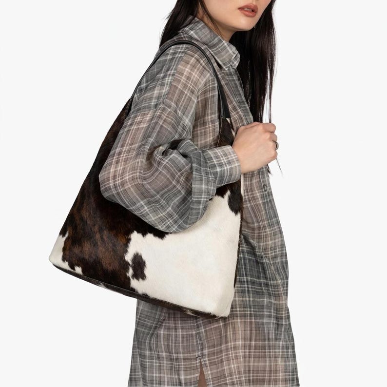 May include: A shoulder bag featuring a cowhide pattern with patches of dark brown and white fur. The bag has a structured silhouette with black leather straps and is worn over the shoulder, showcasing a natural animal print texture throughout.