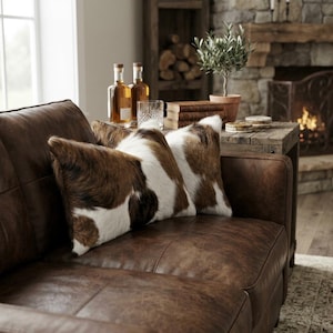 May include: Two rectangular throw pillows featuring a brown and white cowhide pattern with a soft, textured fur appearance. The pillows are positioned on a dark brown leather sofa, showcasing a natural hide aesthetic with irregular patches.
