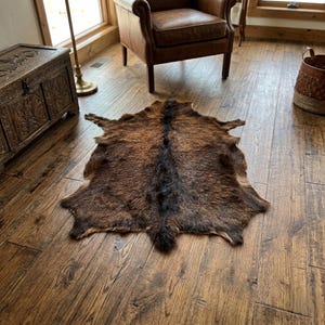 May include: A natural animal hide rug featuring a mottled pattern of dark brown, tan, and black fur. The rug has an irregular, organic shape with a distinct dark stripe running down the centre, showcasing the texture and colour variations of the fur.