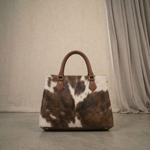 Könnte beinhalten: Eine strukturierte Handtasche mit Kuhfellmuster in dunkelbraunen und weißen Flecken. Die Tasche verfügt über zwei braune Ledergriffe, die mit Metallbeschlägen befestigt sind, sowie einen flachen Boden und ein markantes Tiermuster-Design.