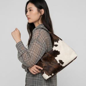 May include: A hobo-style shoulder bag featuring a cowhide pattern with irregular patches of dark brown and white fur. The bag has a curved top silhouette, a single integrated shoulder strap, and dark brown leather trim along the edges.