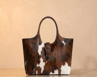 May include: A cowhide tote bag in brown and white, featuring a leather handle and buckle fastening. The bag has a rectangular form with a structured design. The handle and trim are a darker brown, contrasting with the lighter cowhide pattern.