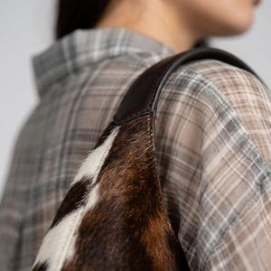 May include: A shoulder bag crafted from brown and white cowhide with a textured, hairy surface. The bag features a smooth, dark brown leather strap attached to the top edge, showcasing a distinct contrast between the fur and the leather material.