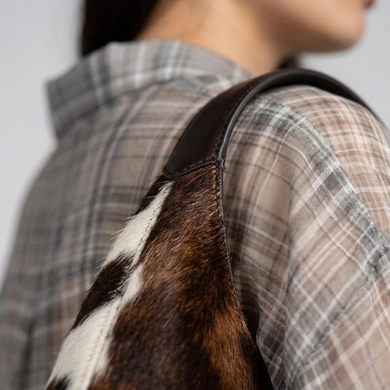 May include: A shoulder bag crafted from brown and white cowhide with a textured, hairy surface. The bag features a smooth, dark brown leather strap attached to the top edge, showcasing a distinct contrast between the fur and the leather material.