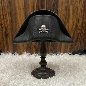 May include: A black leather tricorn hat featuring a silver-toned skull and crossbones emblem centred on the front. The hat is displayed on a dark wooden stand, showcasing the textured material and the distinct three-cornered shape of the headwear.