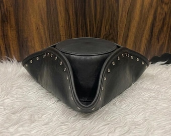 May include: A black leather tricorn hat featuring a rounded crown and a wide, upturned brim. The edges of the brim are accented with a series of small, evenly spaced silver-toned metal studs that follow the curved silhouette of the hat.