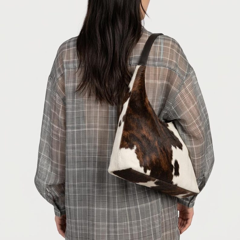 May include: A hobo-style shoulder bag featuring a cowhide pattern with patches of dark brown and white fur. The bag has a single dark brown leather strap and a triangular silhouette, worn over the shoulder against a grey plaid button-down shirt.