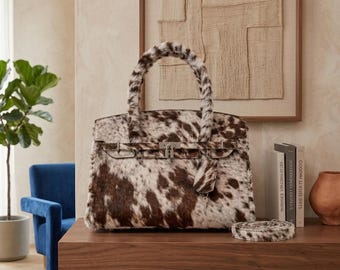 May include: A structured handbag featuring a brown and white cowhide pattern with a matching fur-textured handle. The bag includes a silver-toned metal lock closure on the front, a hanging leather tag, and a detachable matching shoulder strap.