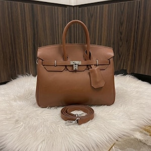 May include: A structured brown leather handbag featuring dual top handles, a front flap with a silver-toned metal padlock closure, a matching leather clochette, and a detachable shoulder strap with silver-toned metal lobster claw clasps.