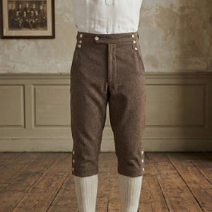 Puede incluir: Pantalones marrones hasta la rodilla con diseño de cintura alta y cierre de botones frontal. Los pantalones incluyen dos bolsillos laterales, cada uno decorado con dos pequeños botones, y tres botones en el borde exterior de cada dobladillo.