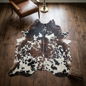 May include: A cowhide rug with a natural pattern of brown, black, and white markings. The rug is irregularly shaped and lies on a dark wood floor. A brown leather chair and a gold lamp are in the background.